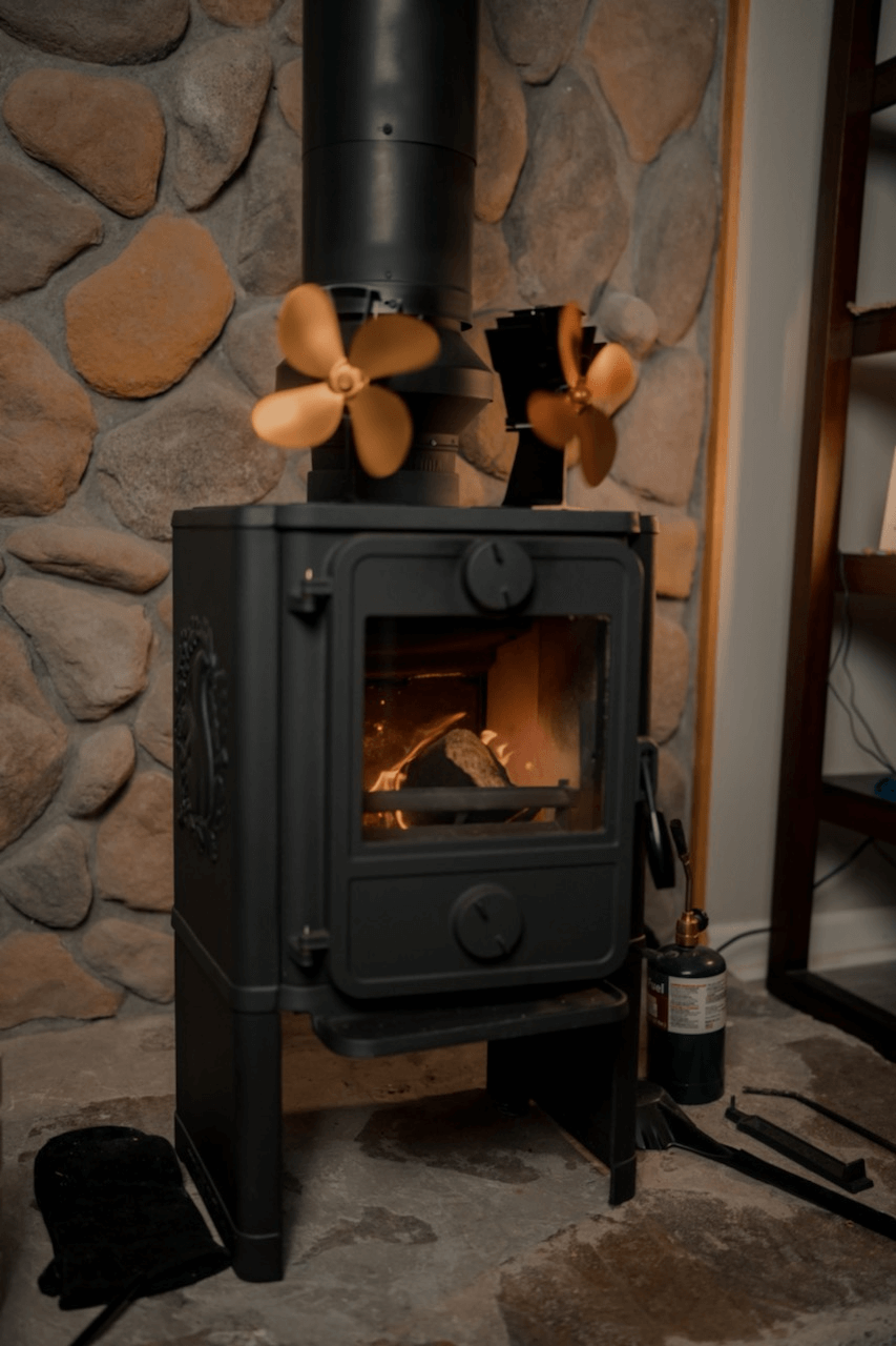 wood burning stove in front of rock wall with 2 gold thermal electric fans on top.
